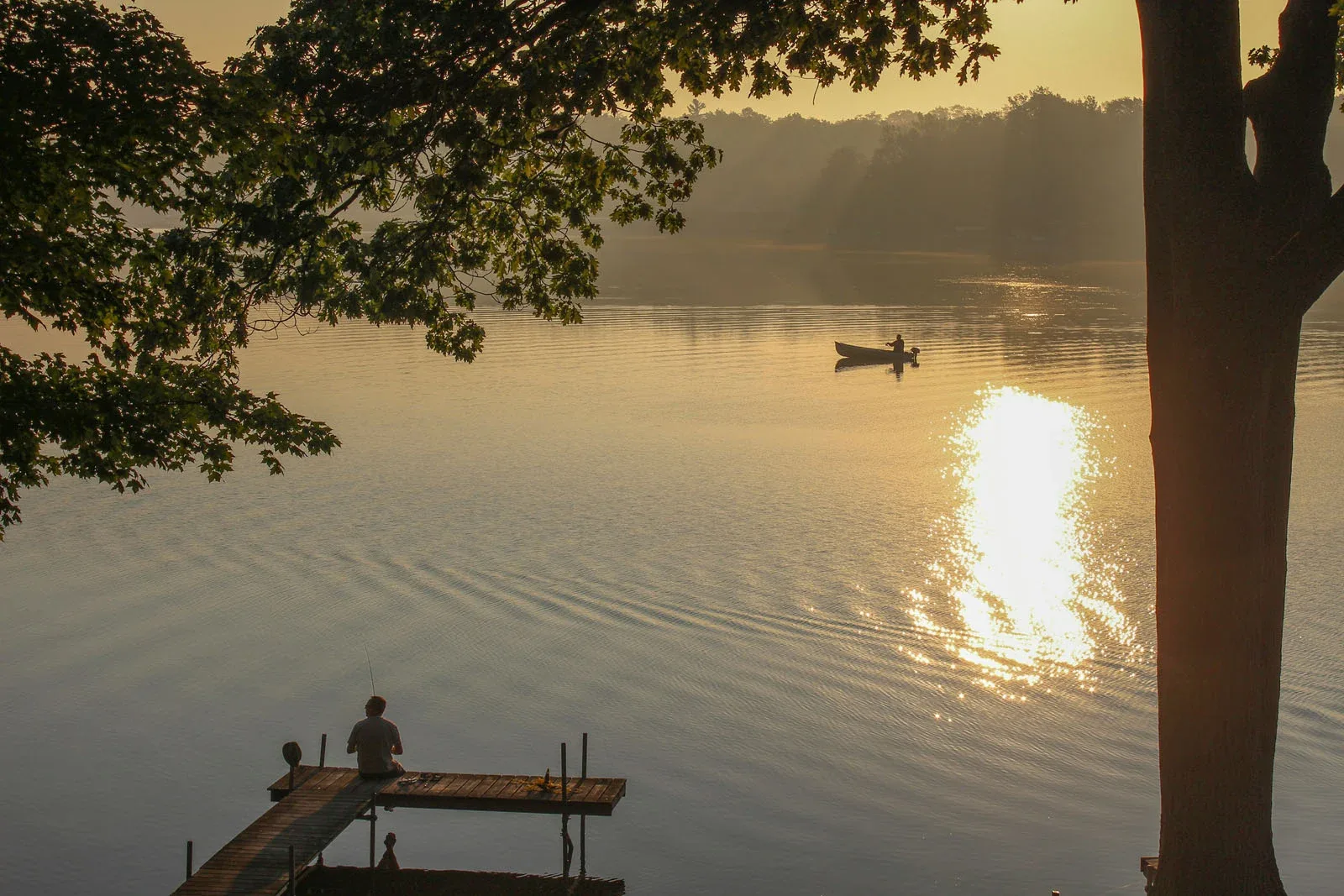 A person sits on a wooden dock fishing in a calm lake at sunrise, while another person in a small boat floats nearby.