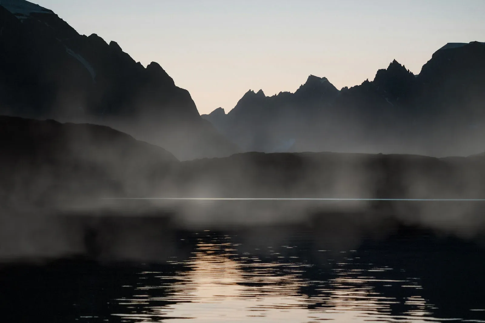 Jagged mountain silhouettes rise above a calm body of water shrouded in low-hanging mist at twilight.