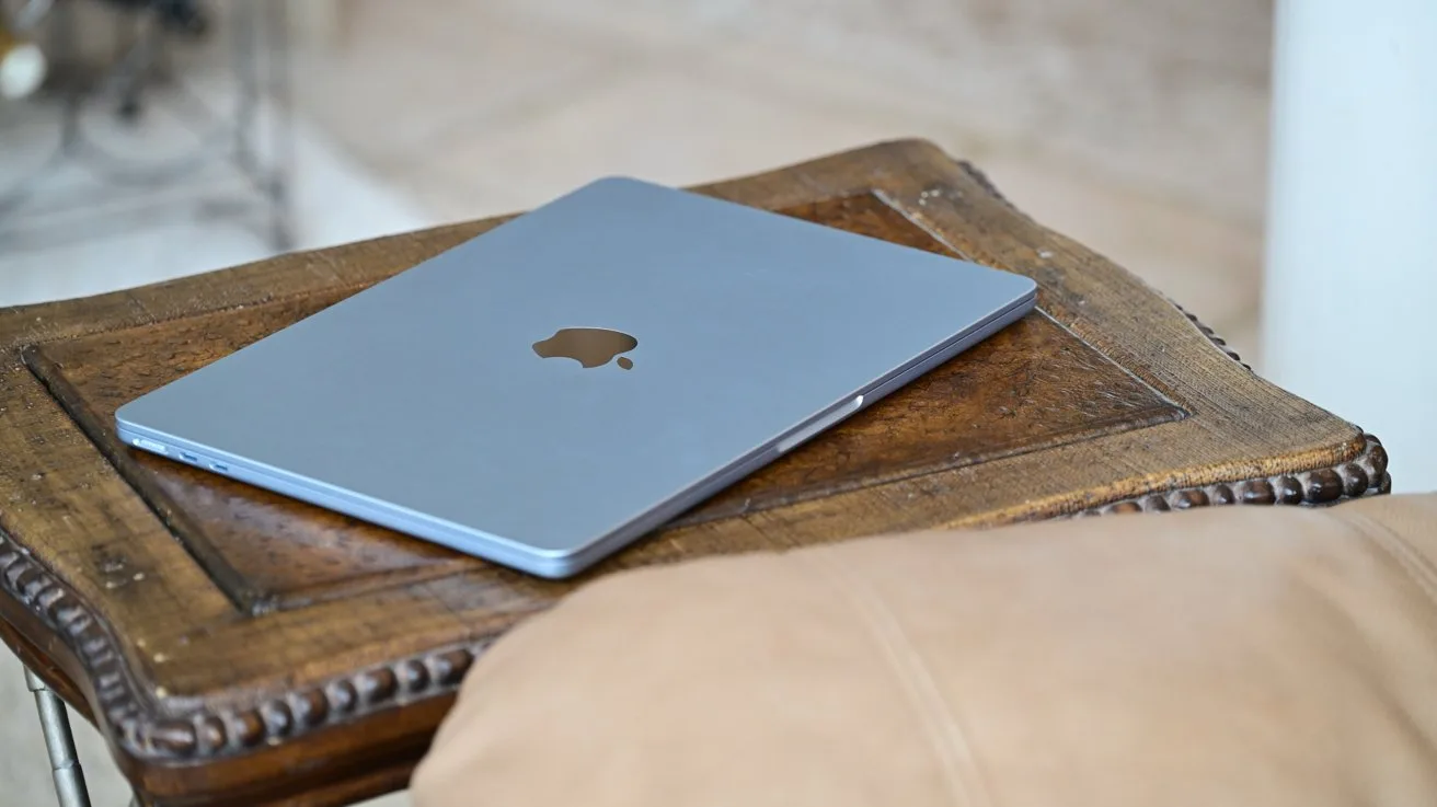 Closed silver MacBook lying on a rustic wooden table beside a tan cushion in a softly lit room, emphasizing a relaxed, homey workspace atmosphere