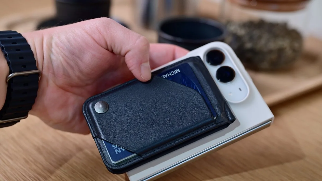 Hand holding a slim black wallet with visible card, placing it onto the back of a white iPhone Duo with dual cameras on a wooden table indoors