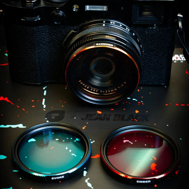A black camera with a 23mm lens sits above two circular camera filters, one teal labeled "EVOKE" and one red labeled "EMBER," resting on a black surface with colorful paint splatters.