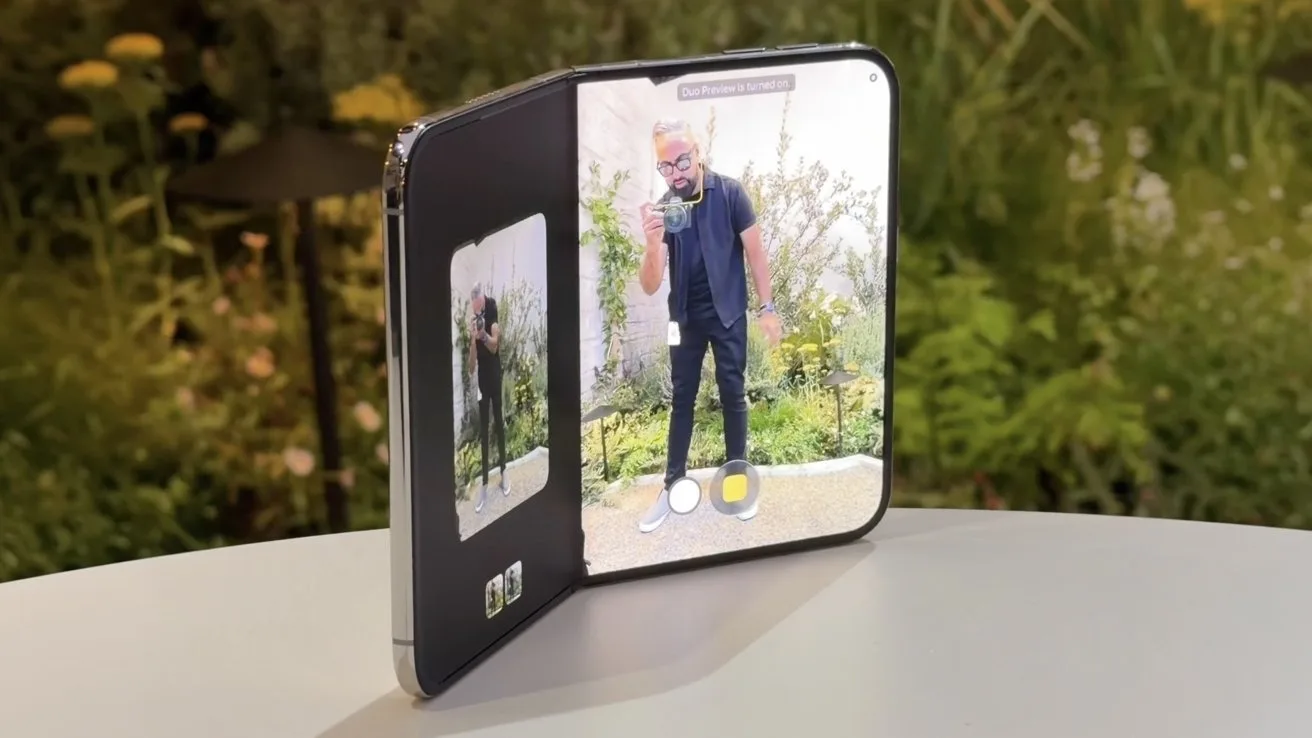 Foldable smartphone partially open on a table, using its large inner screen as a camera viewfinder showing a man posing outdoors, with greenery background and smaller preview display on the left