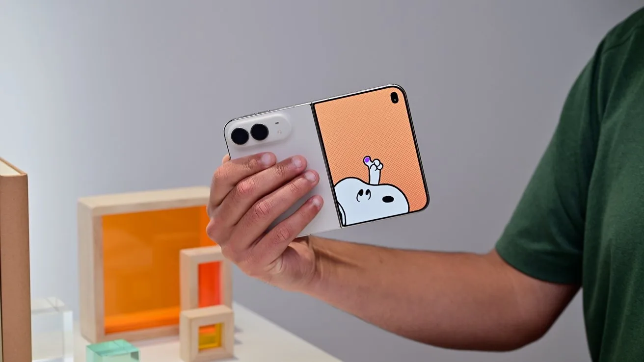 Person holding a foldable smartphone with a playful cartoon character on the unfolded screen, standing indoors near colorful geometric decor blocks and a plain light-colored wall