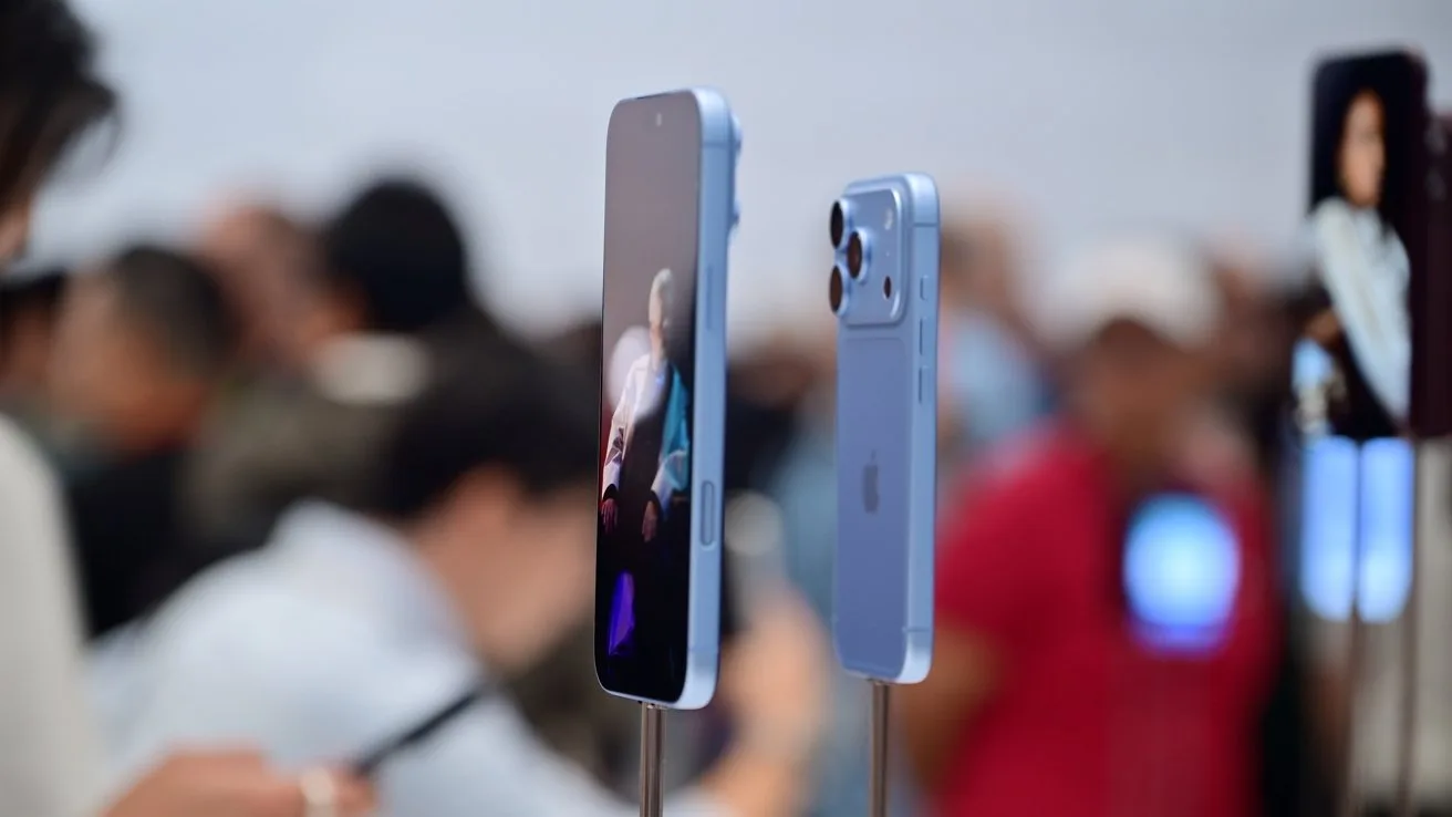 Light blue smartphones on display stands at a busy tech event, screens lit and backs visible, with a blurred crowd of people and lights in the background