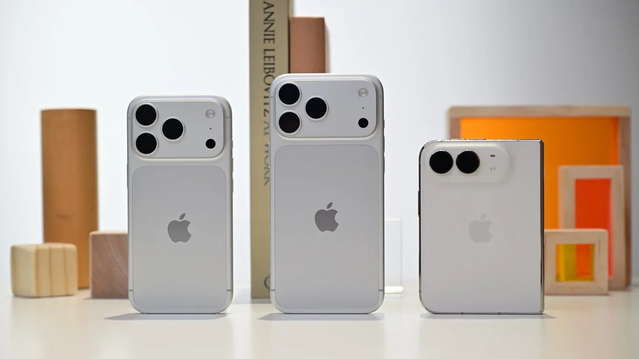 Three silver iPhones with large rear cameras stand upright on a white surface, surrounded by minimal wooden blocks and colorful geometric frames in a softly lit, modern setting