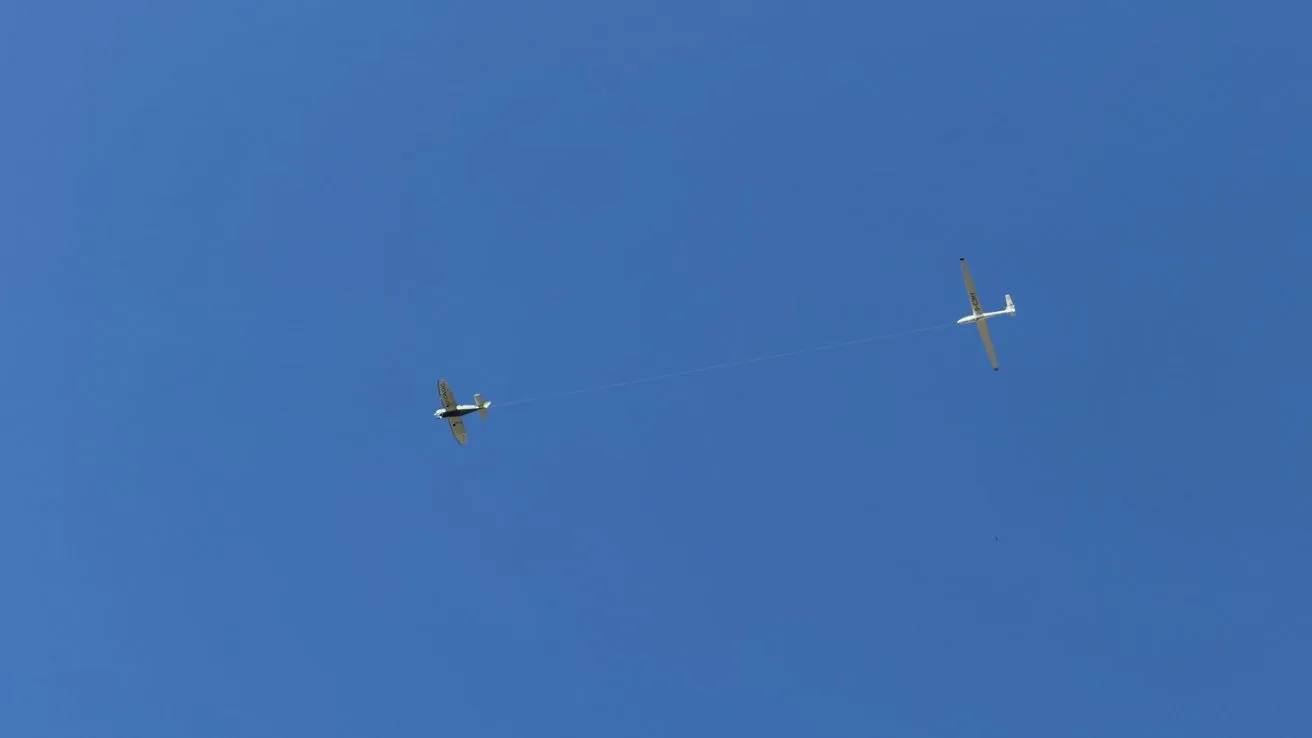 Small airplane towing a white glider with a long cable across a clear blue sky, both aircraft seen from below with wings outstretched and no clouds visible