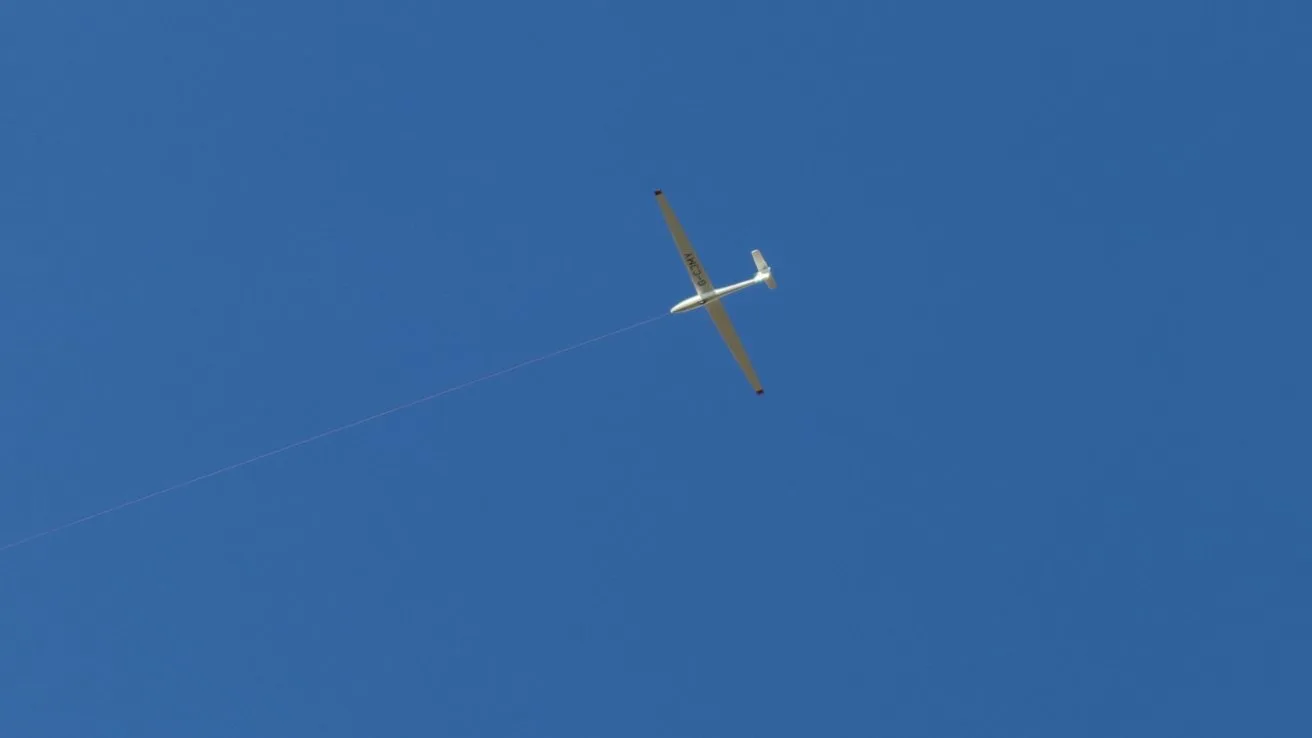 White glider aircraft seen from below against a clear blue sky, with a faint towline trailing behind it