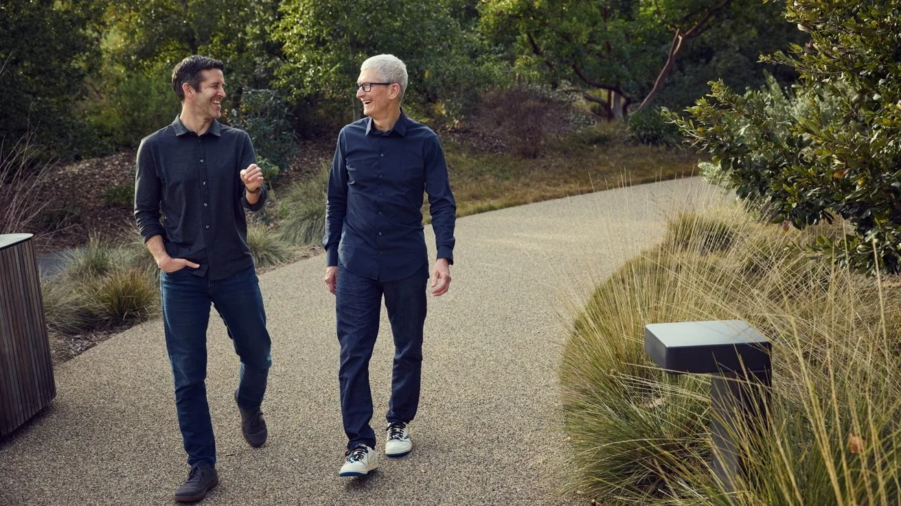 Two men in casual dark clothing walk and talk along a curving paved path in a landscaped park, surrounded by trees, shrubs, and tall grasses on a sunny day
