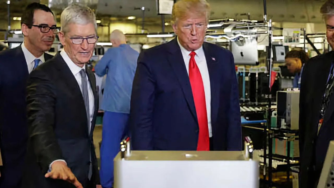 Several men in business suits tour a busy electronics factory, examining a large metallic device while workers and computer equipment appear in the background