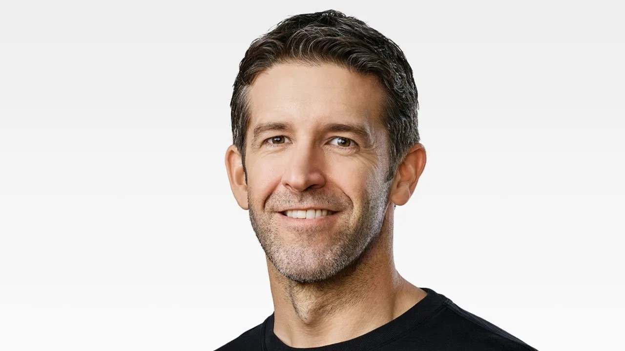 Middle-aged man with short dark hair, light stubble, and a friendly smile, wearing a black crew-neck shirt against a plain light gray background