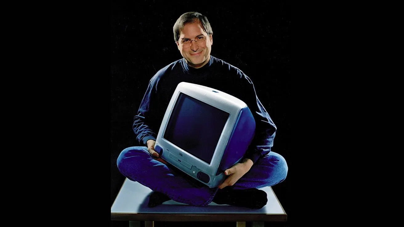 Smiling man in glasses and dark turtleneck sits cross-legged on a table, holding a colorful translucent CRT desktop computer against a dark background