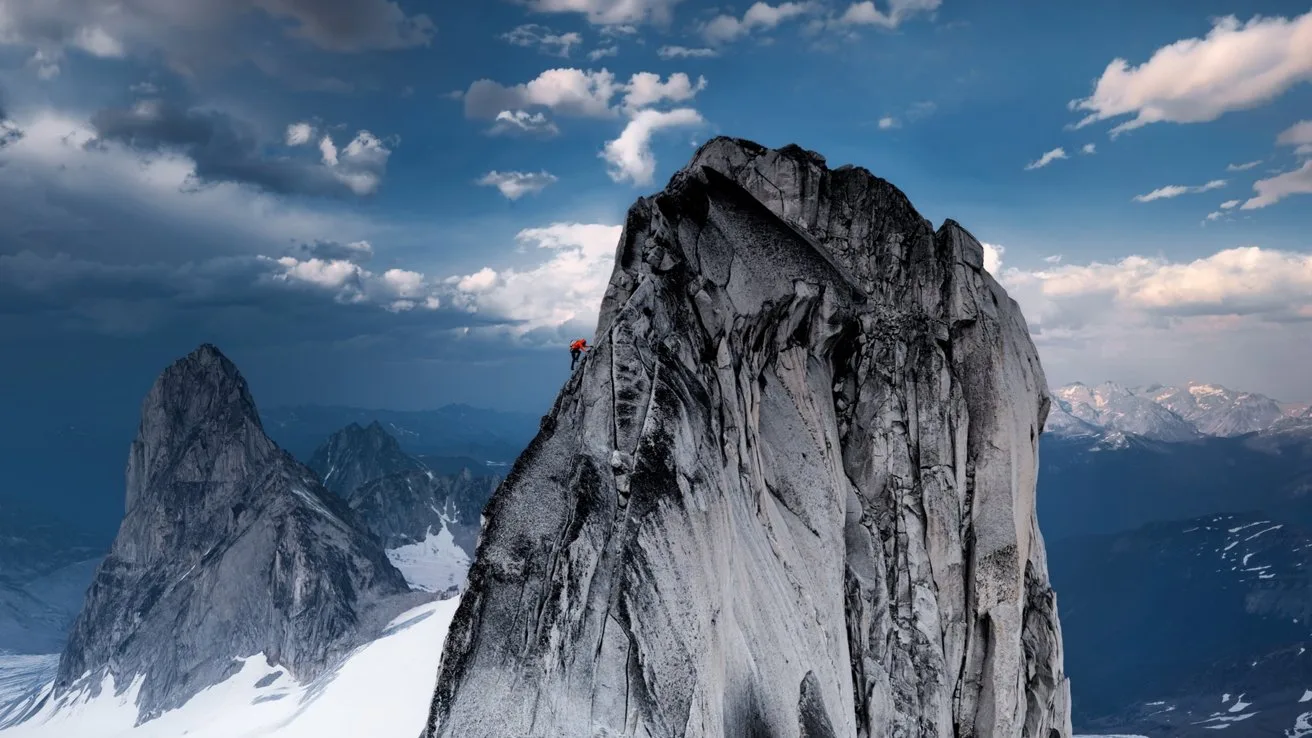 Steep jagged granite peak rising above snowy slopes, with a tiny climber in red near the summit, dramatic clouds and distant mountain range under a deep blue sky