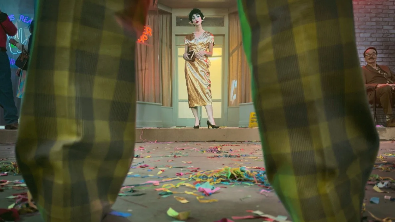 Confetti-strewn floor at a party, viewed between large plaid-clad legs, with a woman in a yellow dress standing center stage under bright lights while seated guests watch in the background