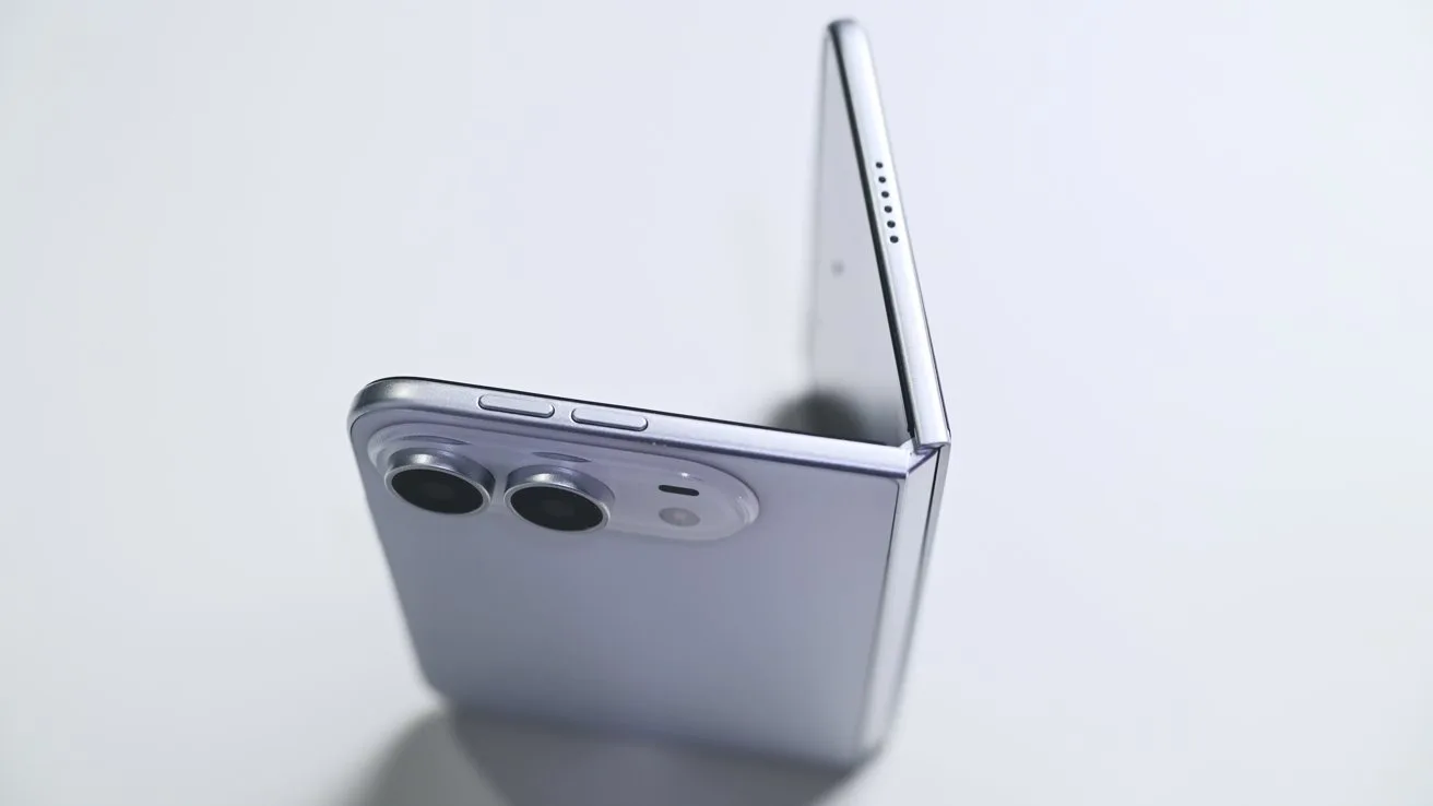 Silver foldable smartphone partially open on a light surface, showing dual rear cameras, side buttons, thin hinge, and upper speaker holes in a clean, minimalist setting