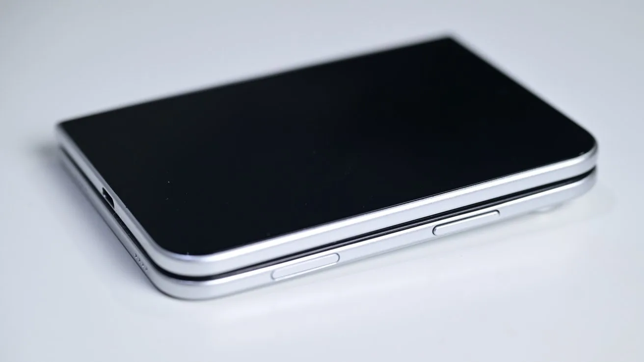 Closed silver and black foldable smartphone lying flat on a white surface, showing its side buttons and slim hinge edge