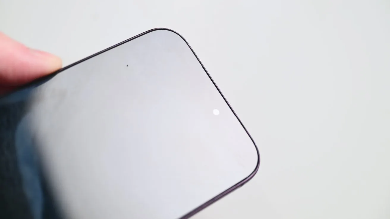 Close-up of a smartphone's top corner with the front camera and sensor barely visible on a reflective screen, held against a plain light background