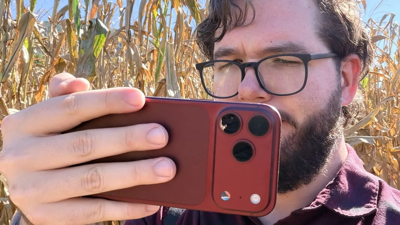 Bearded man with glasses holding a red smartphone horizontally, taking a photo or video, standing in front of tall dry corn stalks on a sunny day