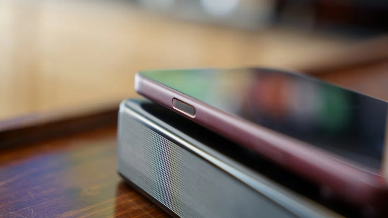 iPhone 18 Pro Max resting on top of a rectangular speaker on a wooden surface, focused on the phone's side button with a softly blurred background