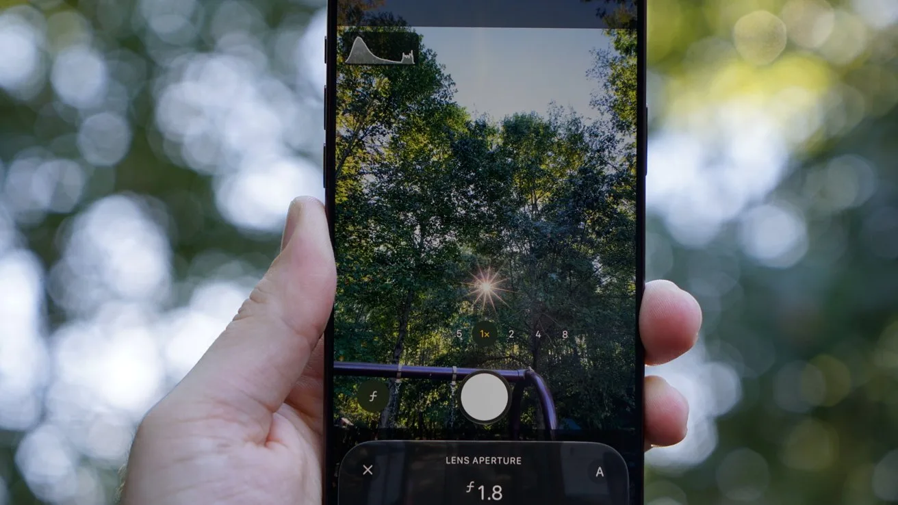 Hand holding an iPhone 18 Pro Max outdoors, camera app open, focusing on sunlit green tree canopy with bright sunburst shining through leaves, blurred leafy background surrounding the phone