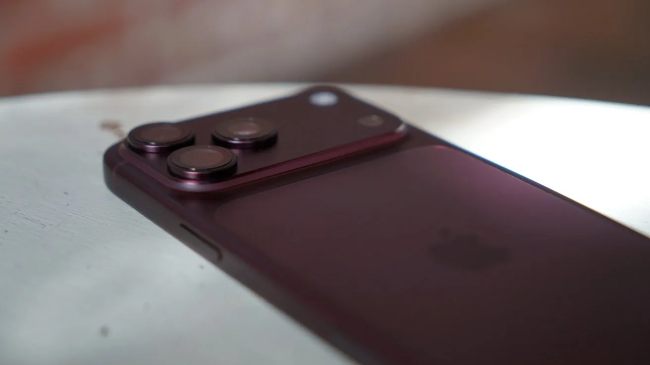 Burgundy iPhone 18 Pro Max lying on a light surface, showing raised triple rear camera lenses and side buttons, with soft natural light highlighting the sleek metallic finish