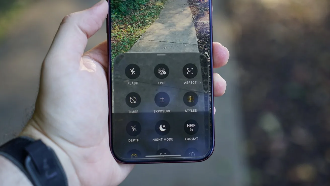 Hand holding an iPhone 18 Pro Max outdoors, camera app open displaying advanced photo controls over a view of a sidewalk path surrounded by grass and blurred trees in the background