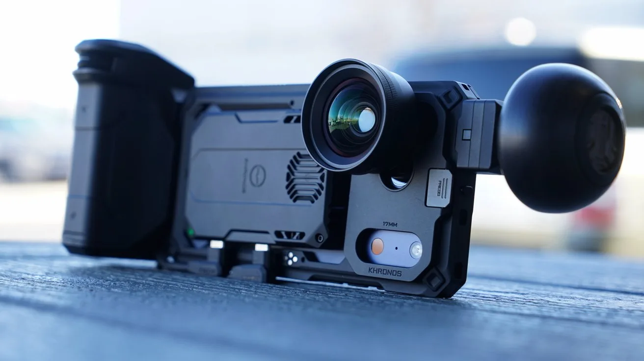 iPhone mounted in a rugged camera rig with a large external lens and grip, resting on a table outdoors, resembling a compact professional video or photography setup