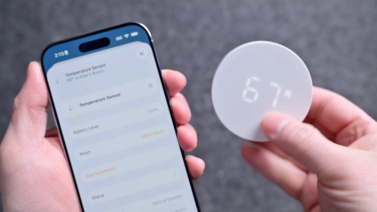 Hands holding a smartphone app and a small round digital temperature sensor displaying 67, showing a connected home device setup against a neutral gray background