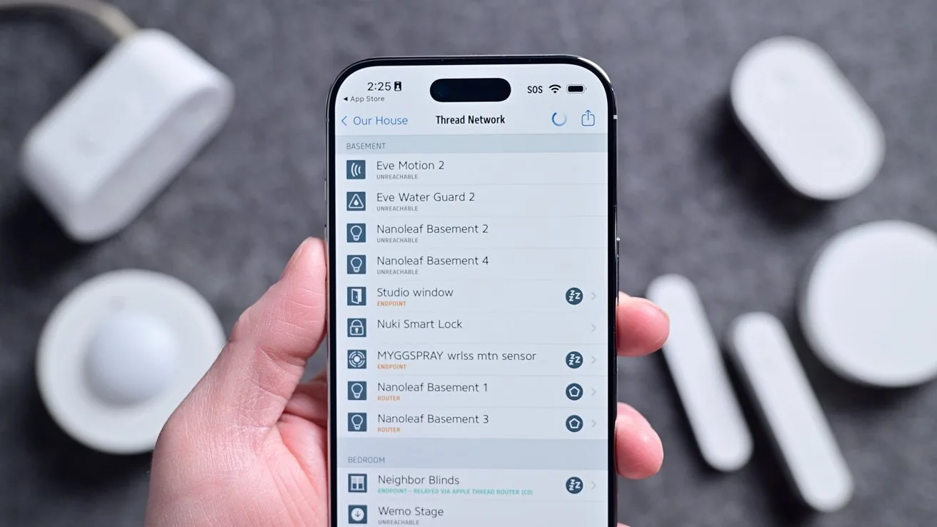 Hand holding a smartphone displaying a smart home app with a list of connected devices, surrounded by several small white smart home sensors and gadgets on a gray surface