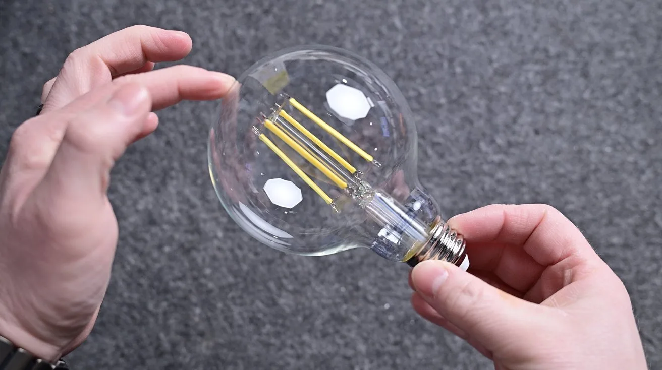 Two hands holding a large clear LED filament light bulb against a gray textured background, with fingers gently touching the glass near the top of the round bulb