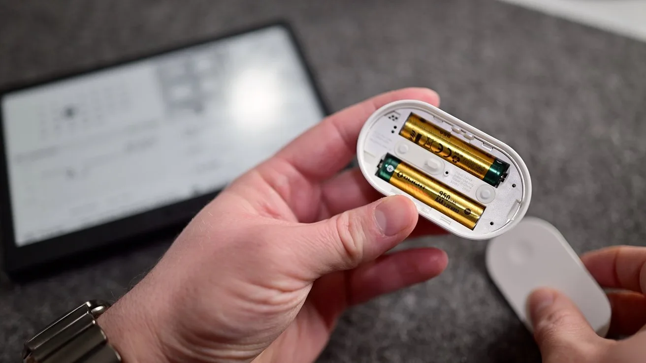Hands holding a small oval device with its battery compartment open, revealing two AA batteries, while a blurred electronic screen or e-reader lies on a gray surface in the background