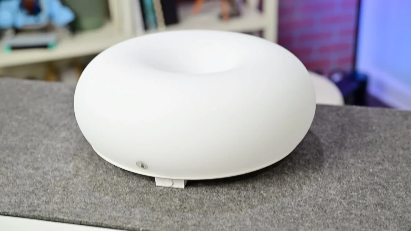 White, donut-shaped electronic device with a smooth matte surface sitting on a gray countertop, with shelves and books softly blurred in the background