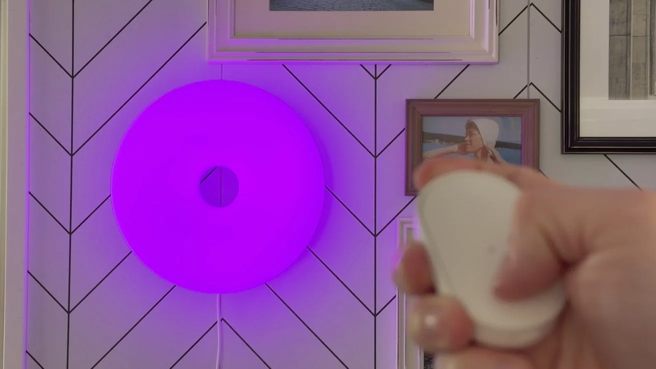 Hand holding a small white remote control in front of a round purple wall light, mounted on a chevron-patterned wall with framed photographs in the background