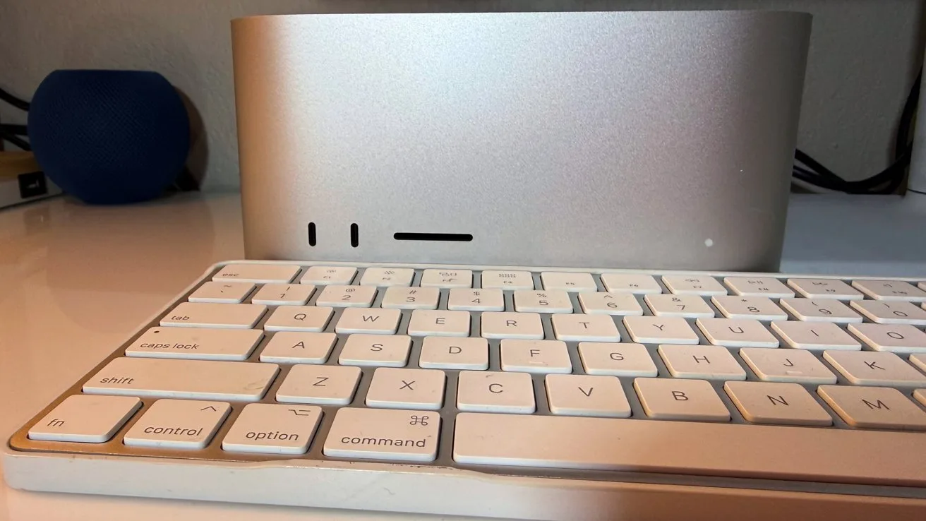 White Apple-style keyboard on a desk in front of a compact silver desktop computer, with ports visible on the front and a small blue speaker in the background