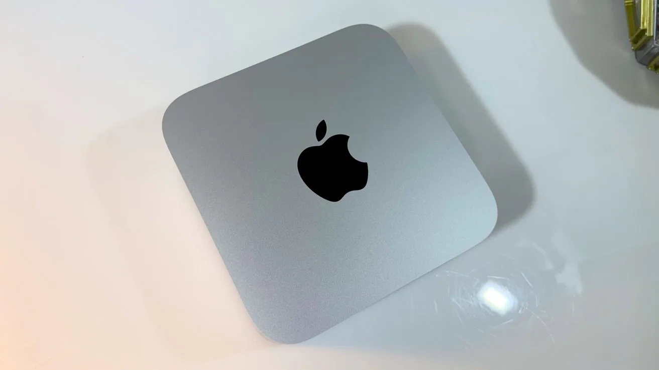 Silver Apple Mac mini computer on a glossy white surface, viewed from above, showing the centered black Apple logo and softly rounded square edges