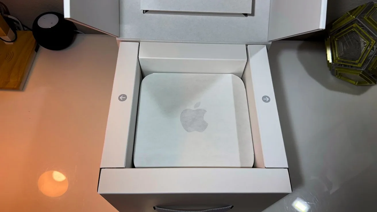 Open white box on a desk containing a new Apple Mac Mini wrapped in translucent protective paper, viewed from above, with soft lighting and a decorative object nearby