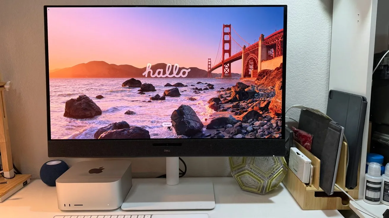 Computer desk setup with large monitor showing Golden Gate Bridge sunset and the word hallo, Mac mini beneath, keyboard, mouse, decorative plant, and organized office supplies nearby