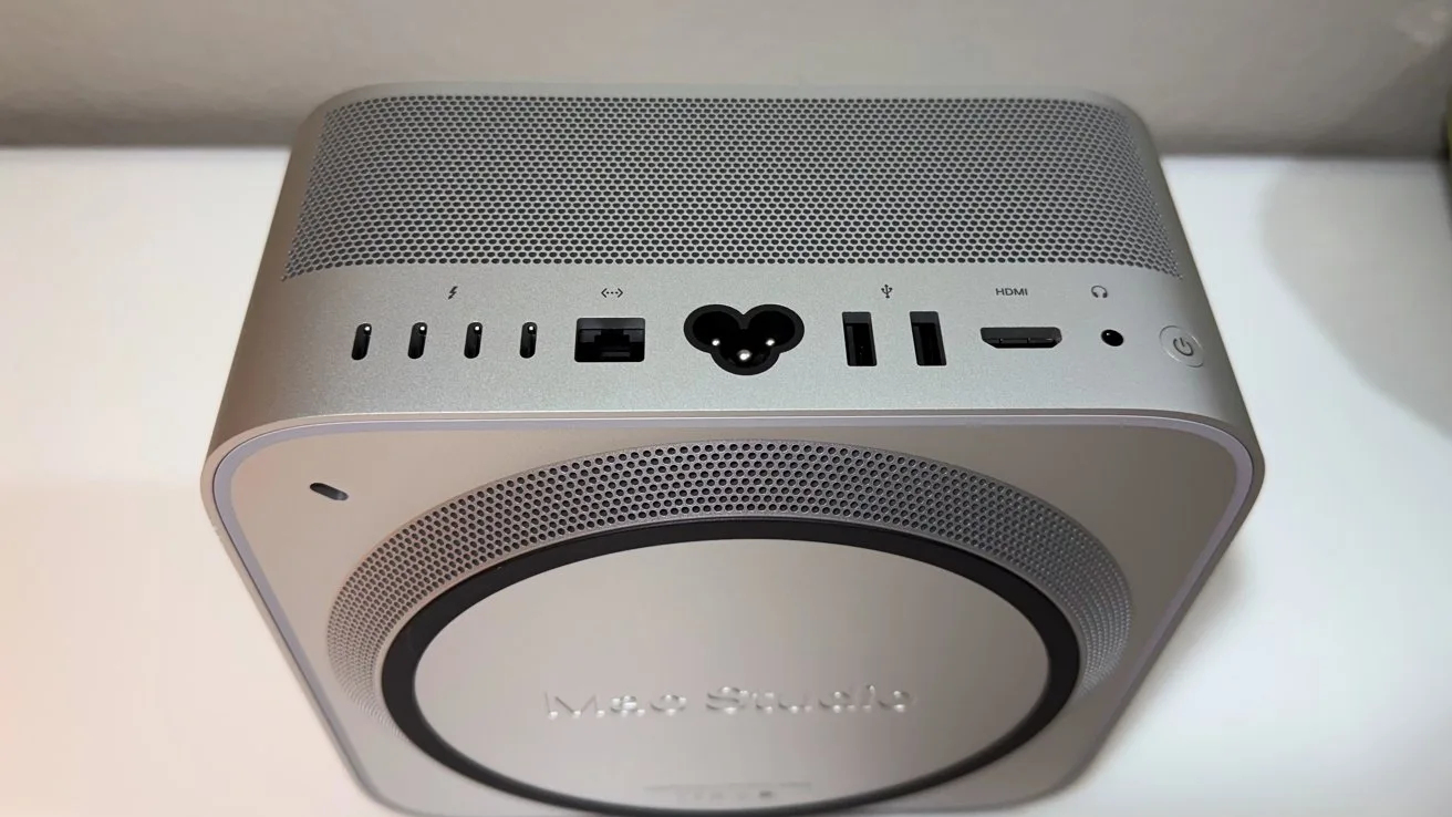 Compact silver desktop computer viewed from above, showing rear ports including power connector, USB, HDMI, Ethernet, and vented speaker-like grille on top, sitting on a white surface