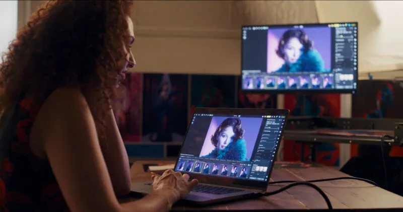 A woman with curly hair sits at a wooden desk, using a laptop to edit a portrait that is also displayed on a larger monitor behind it.