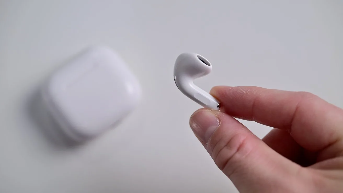 Hand holding a single white wireless earbud in focus, with a blurred white charging case resting on a plain light-colored surface in the background