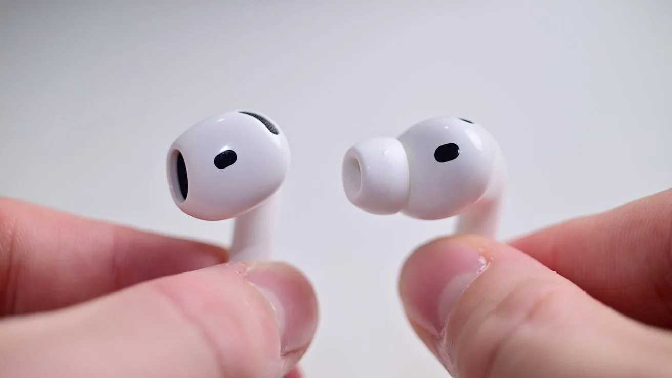 Close-up of two white wireless earbuds with silicone tips, each held between human fingers against a plain light background, showing speaker openings and black vents.