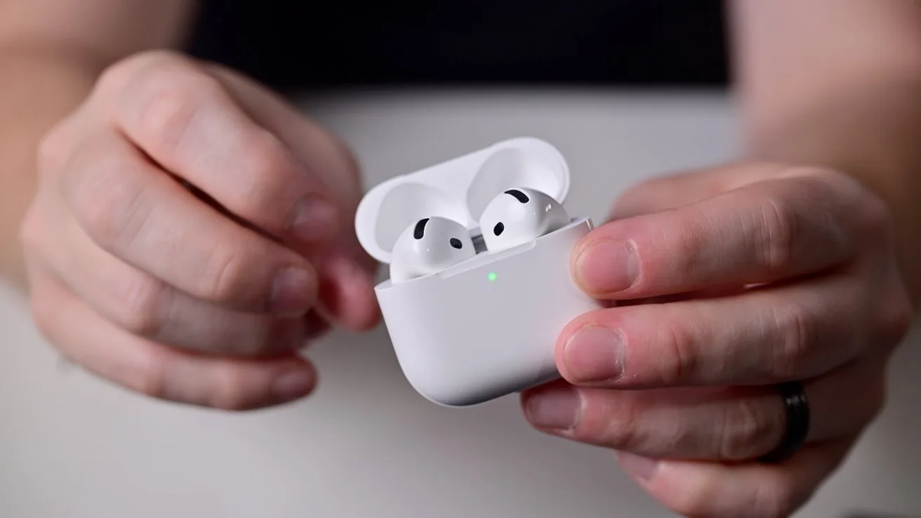 Hands holding an open white wireless earbud case with two earbuds inside, small green charging light on front, against a blurred background
