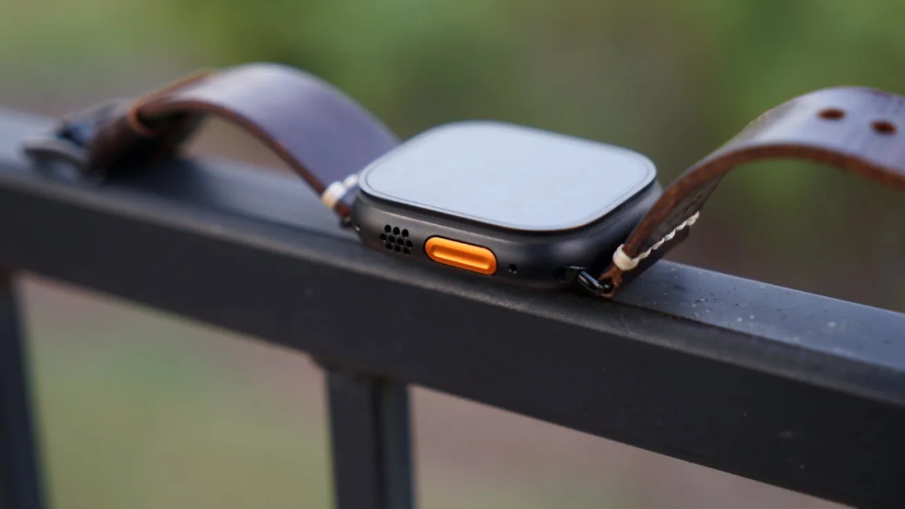 Apple Watch Ultra 4 with brown leather band resting sideways on a black metal railing, showing a square face, speaker holes, and a prominent orange side button against a blurred green background