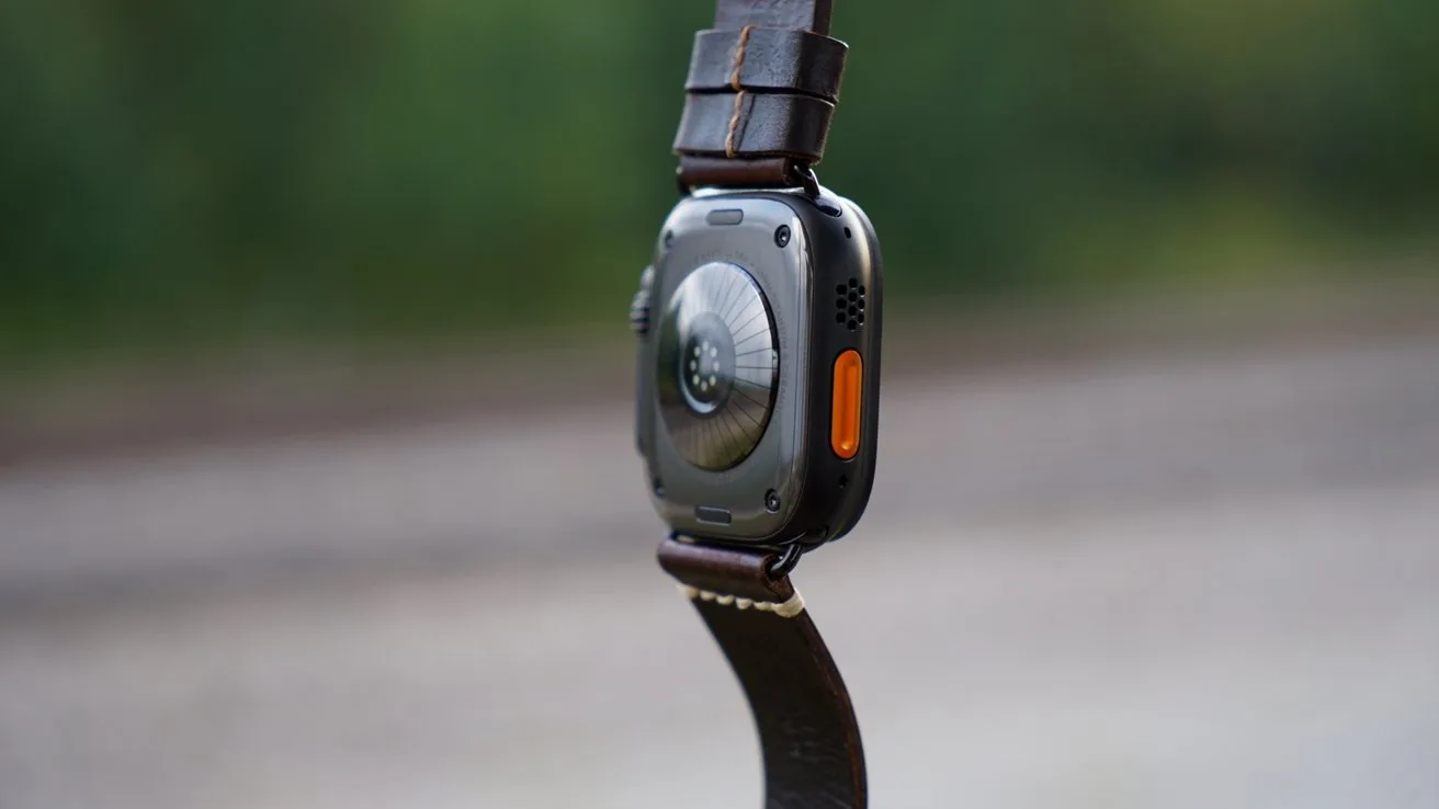 Close-up of a Apple Watch Ultra 4 hanging by its brown leather strap, showing the rounded metal back, sensors, and an orange side button against a blurred outdoor background