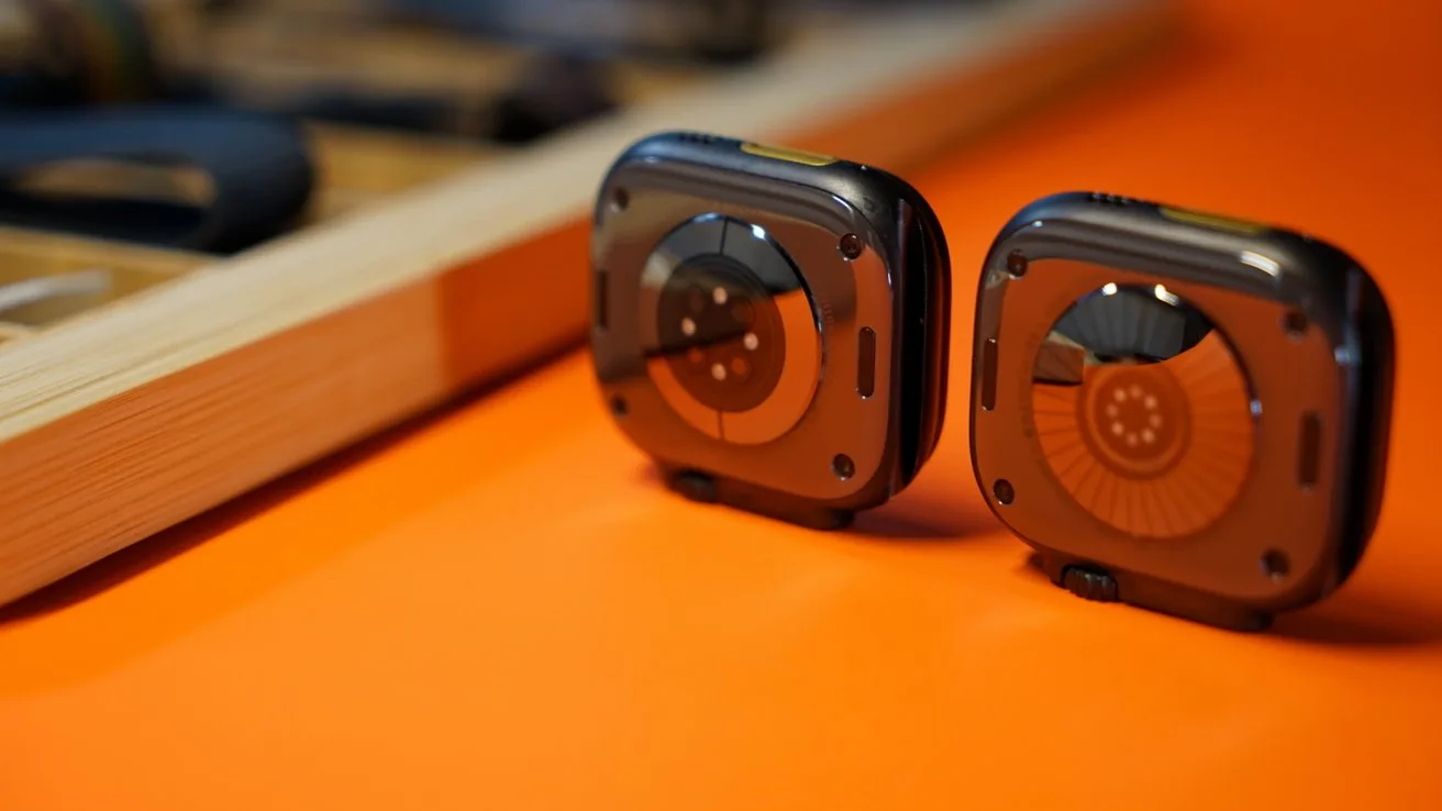 Apple Watch Ultra 3 on the left, Ultra 4 on the right, standing upright on an orange surface, showing their circular sensor backs, with a wooden tray and blurred gadgets in the background