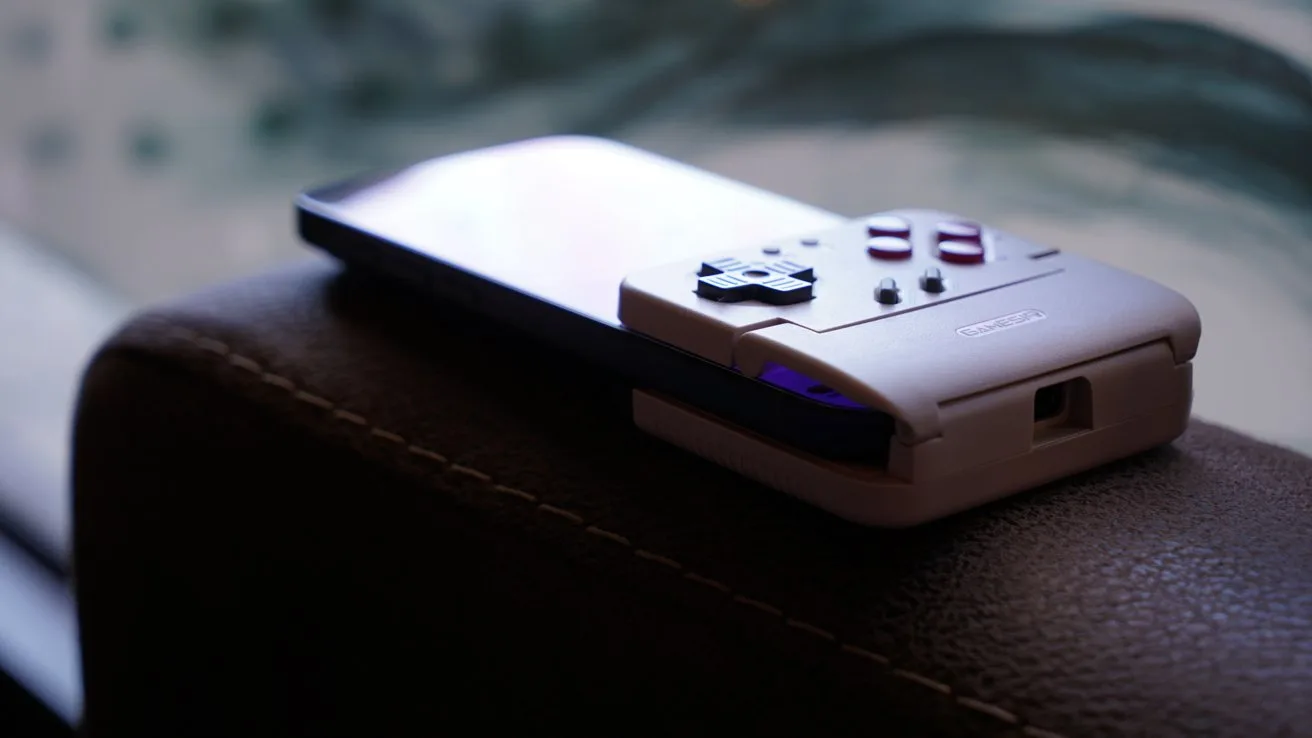 iPhone attached to a retro-style handheld gaming controller with D-pad and buttons, resting on a brown leather surface with soft, blurred background.