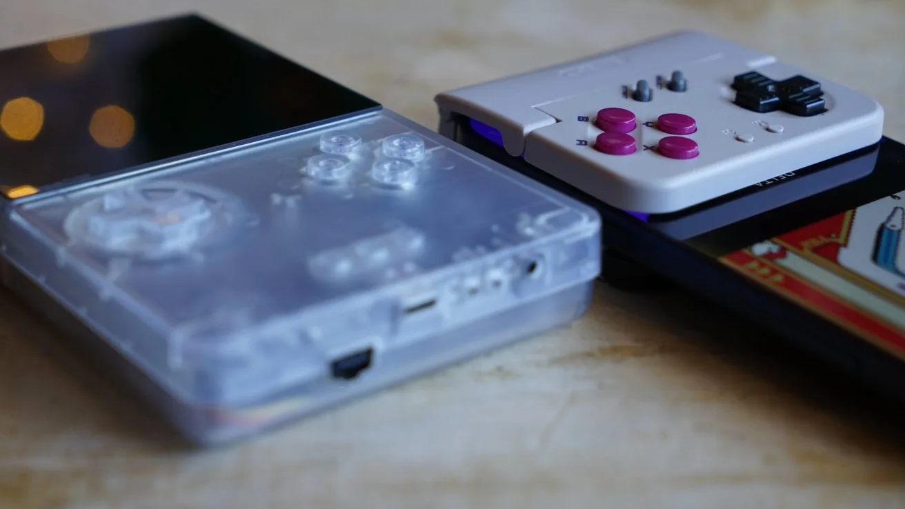 Close-up of a Game Boy-like handheld gaming device and a controller attached to an iPhone on a table, including a transparent controller, a small white console with purple buttons, and a dark device displaying colorful retro graphics