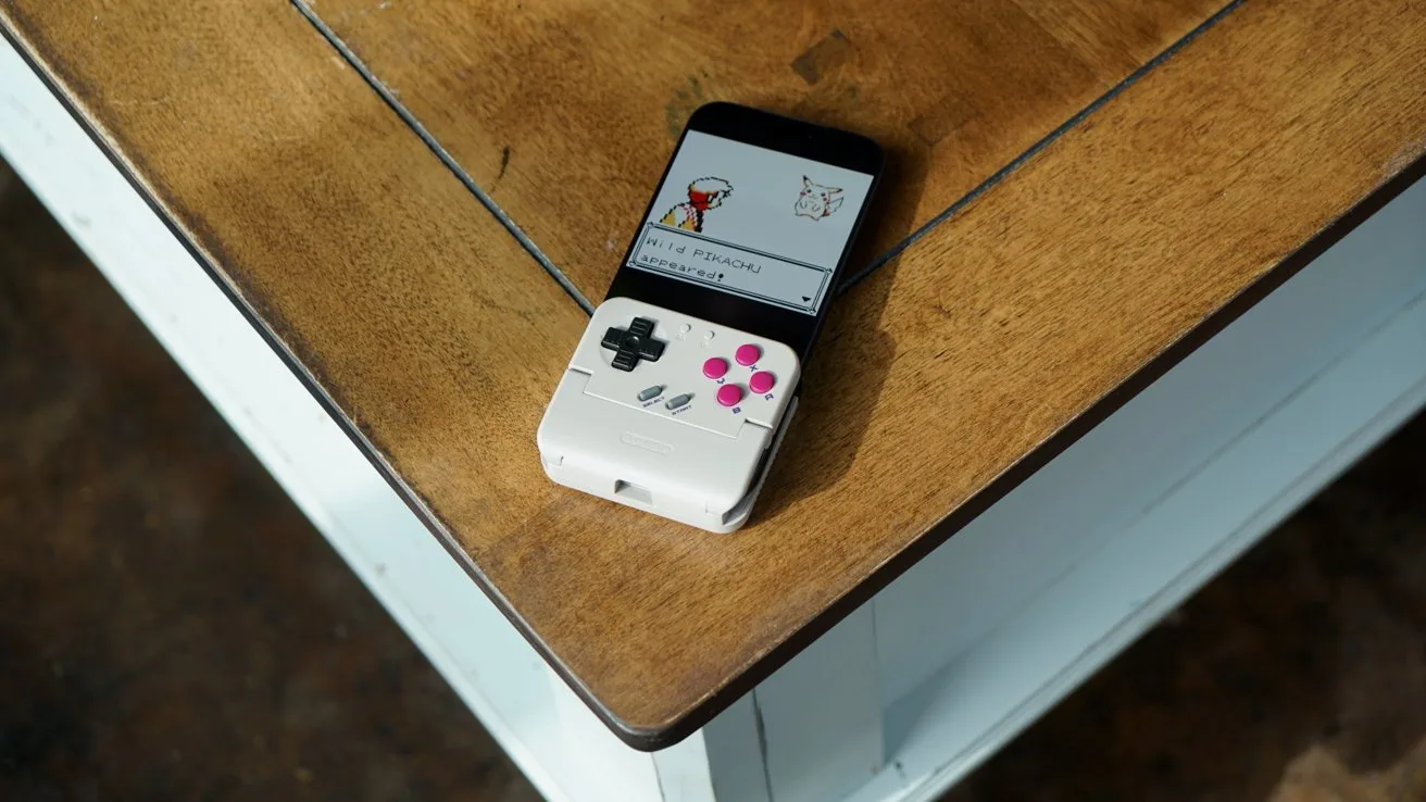 iPhone with attached retro-style game controller showing a Pokemon battle screen, resting on the corner of a wooden table with white sides and a dark floor below