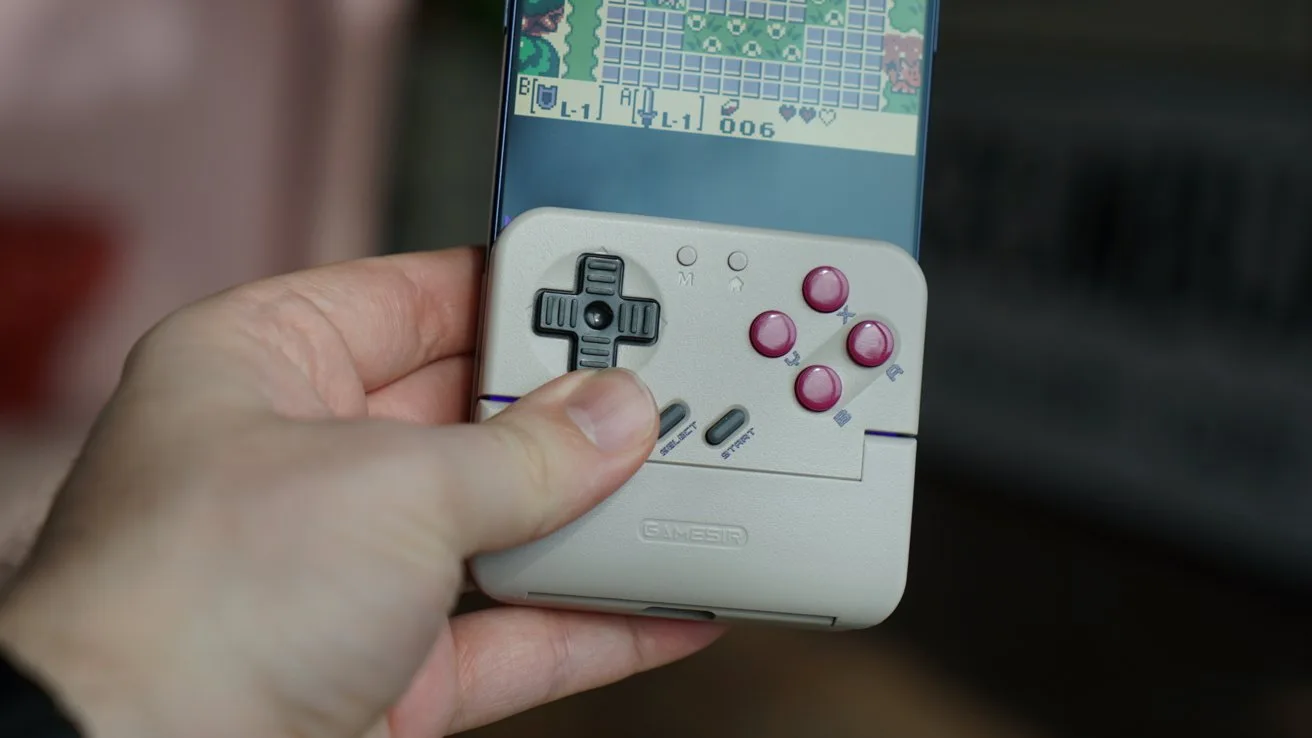 Hand holding an iPhone attached to a small retro-style game controller with gray D-pad, pink buttons, and a pixelated game displayed on the phone screen
