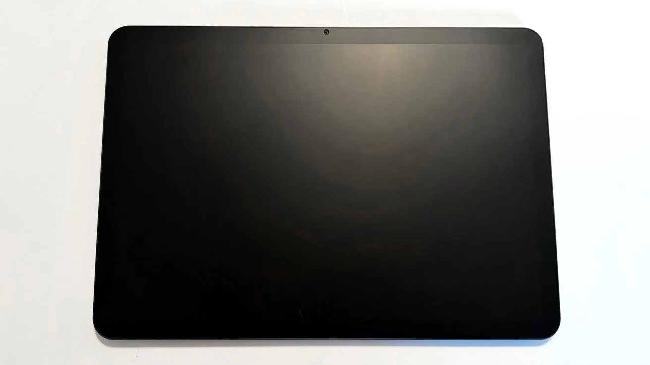 Black tablet computer with a blank dark screen, rounded corners, and a small centered camera at the top, lying flat on a clean white surface