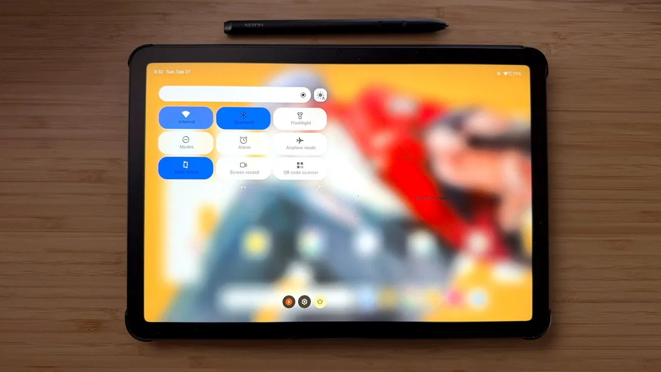 Tablet lying on a wooden surface with a stylus above it, displaying a blurred colorful background and a settings-style control panel with large buttons on the left side
